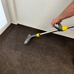 Don Clean - Czyszczenie ciemnobrązowego dywanu za pomocą odkurzacza piorącego z żółtymi elementami w rogu pokoju z białą ścianą i listwami przypodłogowymi.