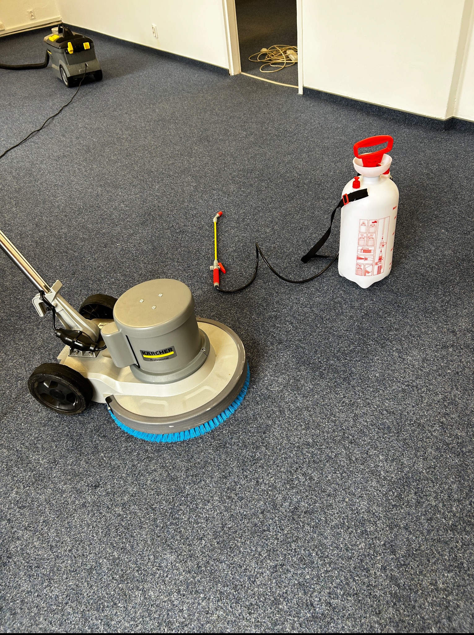 Maszyna Karcher z niebieską szczotką i spryskiwacz na szarym dywanie w trakcie czyszczenia. Widoczny odkurzacz. W tle białe ściany i otwarte drzwi.