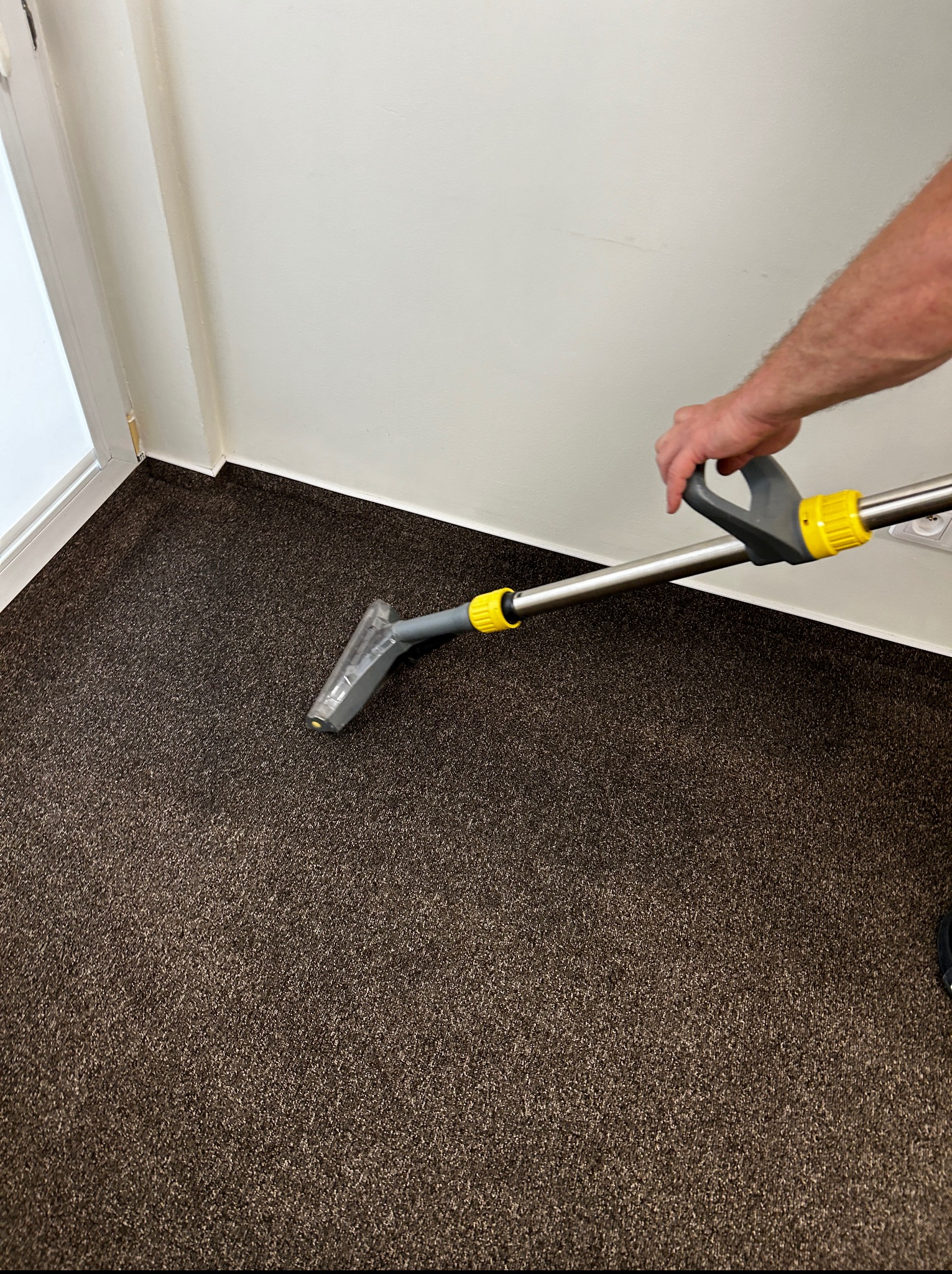 Czyszczenie ciemnobrązowego dywanu za pomocą odkurzacza piorącego z żółtymi elementami w rogu pokoju z białą ścianą i listwami przypodłogowymi.