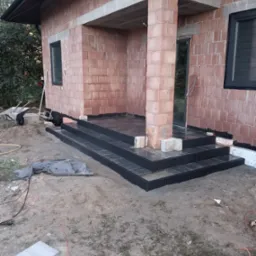 Ciemne schody z kostki prowadzą do surowego budynku z cegły. Widoczne narzędzia i materiały budowlane sugerują etap prac wykończeniowych.