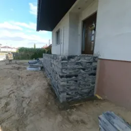 Wykończony cokół budynku z szarej kostki elewacyjnej. Na ziemi leżą materiały budowlane. Widoczny fragment elewacji, drzwi wejściowe i niebo.