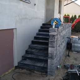 MR. KOP Mateusz Raduj - Zewnętrzne schody z ciemnej kostki, prowadzące do budynku. Widoczna ściana budynku w trakcie wykończenia, kabel zasilający i narzędzia budowlane w tle.