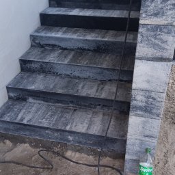 MR. KOP Mateusz Raduj - Ukończone schody kamienne z szarej kostki, z widocznym fragmentem ściany i kablem na ziemi. Butelka Sprite przy podstawie schodów dodaje skali.