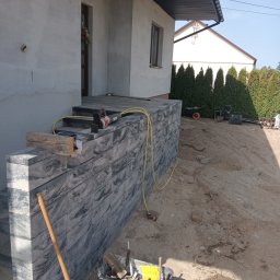 MR. KOP Mateusz Raduj - Wznoszenie schodów wejściowych z szarej kostki, widoczne narzędzia pracy i częściowo ułożona kostka. Prace w toku, dom w tle.