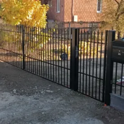 Czarny, metalowy płot z furtką i skrzynką na listy numer 24, na tle domu i drzew z żółtymi liśćmi. Widoczne elementy montażowe i ziemia. Styl nowoczesny.