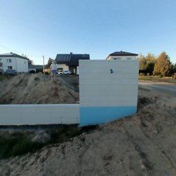 MR. KOP Mateusz Raduj - Fundament pod ogrodzenie panelowe z widocznym wykopem ziemi i osobą na górze. Tło stanowią domy i drzewa w słoneczny dzień.