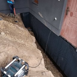 MR. KOP Mateusz Raduj - Wykop fundamentowy z izolacją pionową przy ścianie budynku, pracownik z łopatą w tle, oświetlenie budowlane na pierwszym planie.