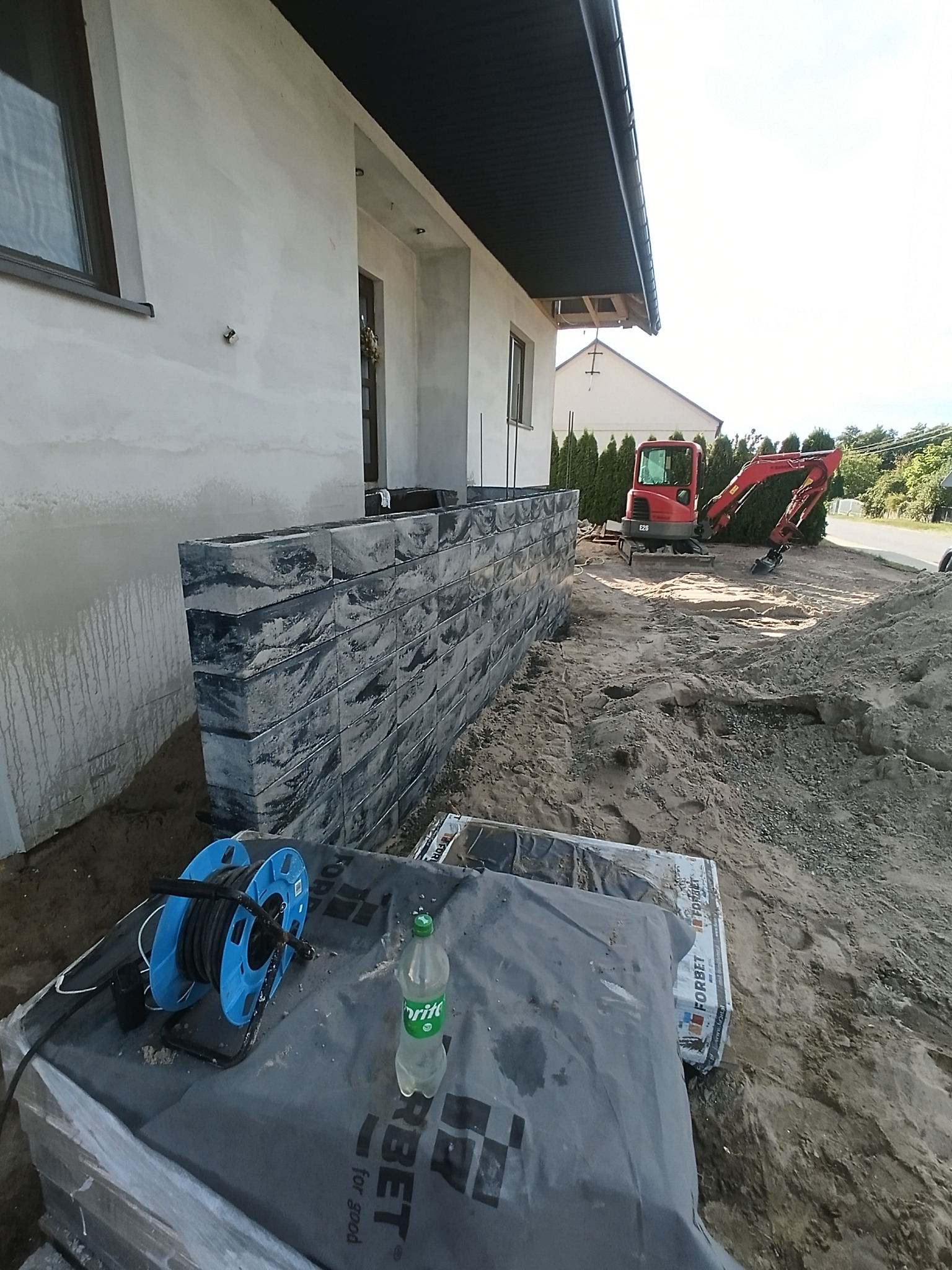 Wznoszenie murku z bloczków betonowych przy budynku. W tle mini koparka i teren budowy. Widoczne materiały budowlane i narzędzia.