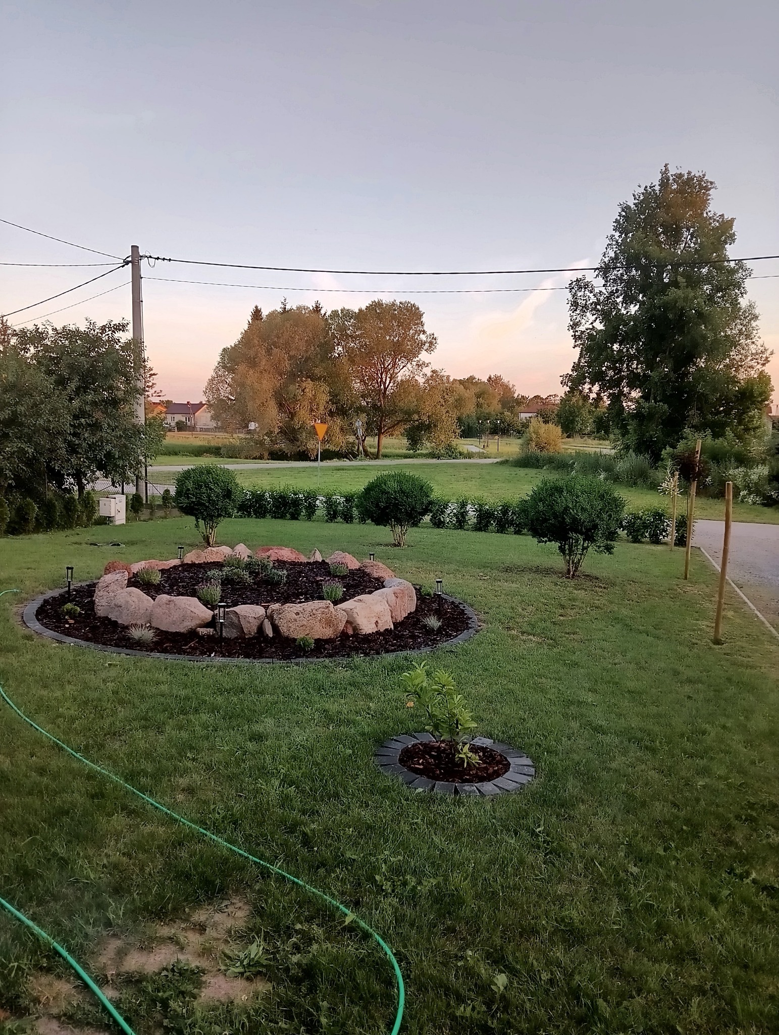 Ogród z okrągłą rabatą otoczoną kamieniami i oświetleniem solarnym, mała rabata z sadzonką, trawnik i drzewa w tle, widok na posesję.