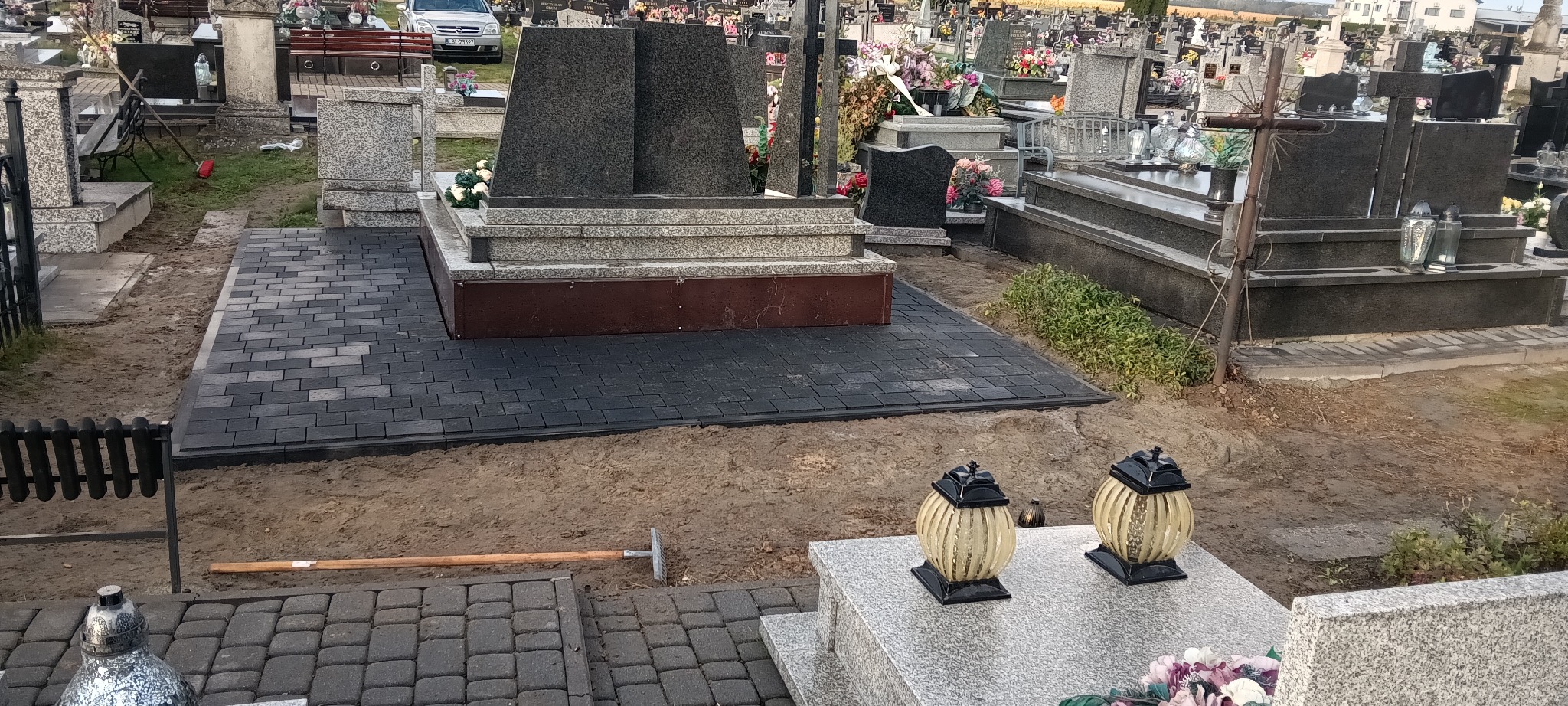 Ciemna kostka brukowa ułożona wokół nagrobka na cmentarzu. Widoczne narzędzia i inne groby w tle. Częściowo widoczne lampiony.