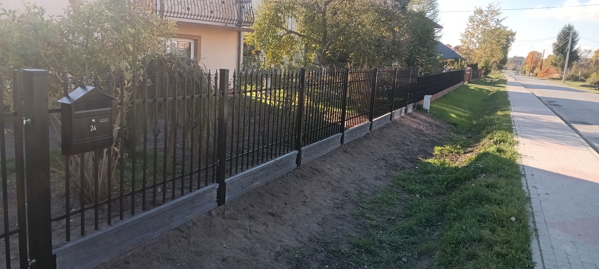 Czarne ogrodzenie panelowe z metalowymi prętami i betonową podmurówką, widok z boku, skrzynka pocztowa z numerem 24 na słupku, trawa i chodnik wzdłuż ulicy.