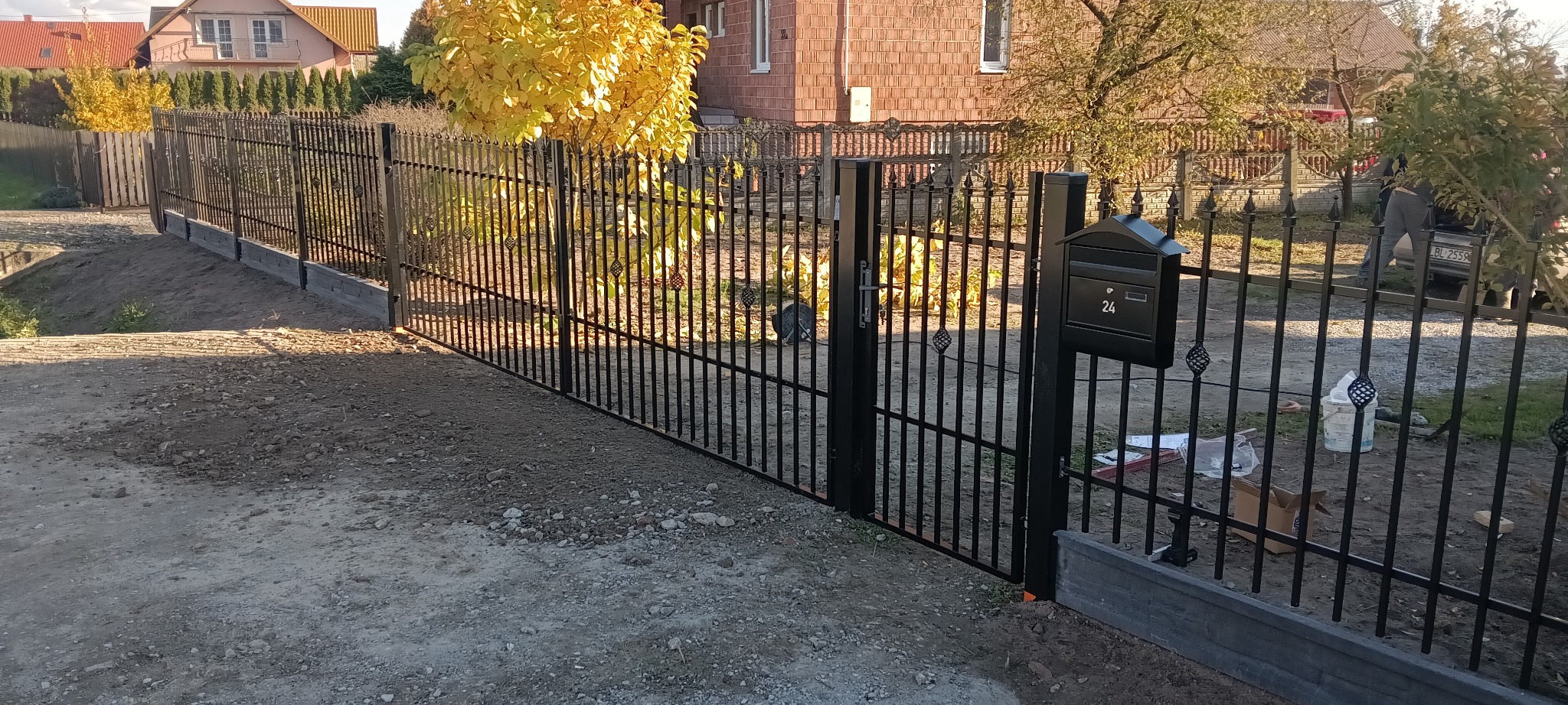 Czarny, metalowy płot z furtką i skrzynką na listy numer 24, na tle domu i drzew z żółtymi liśćmi. Widoczne elementy montażowe i ziemia. Styl nowoczesny.