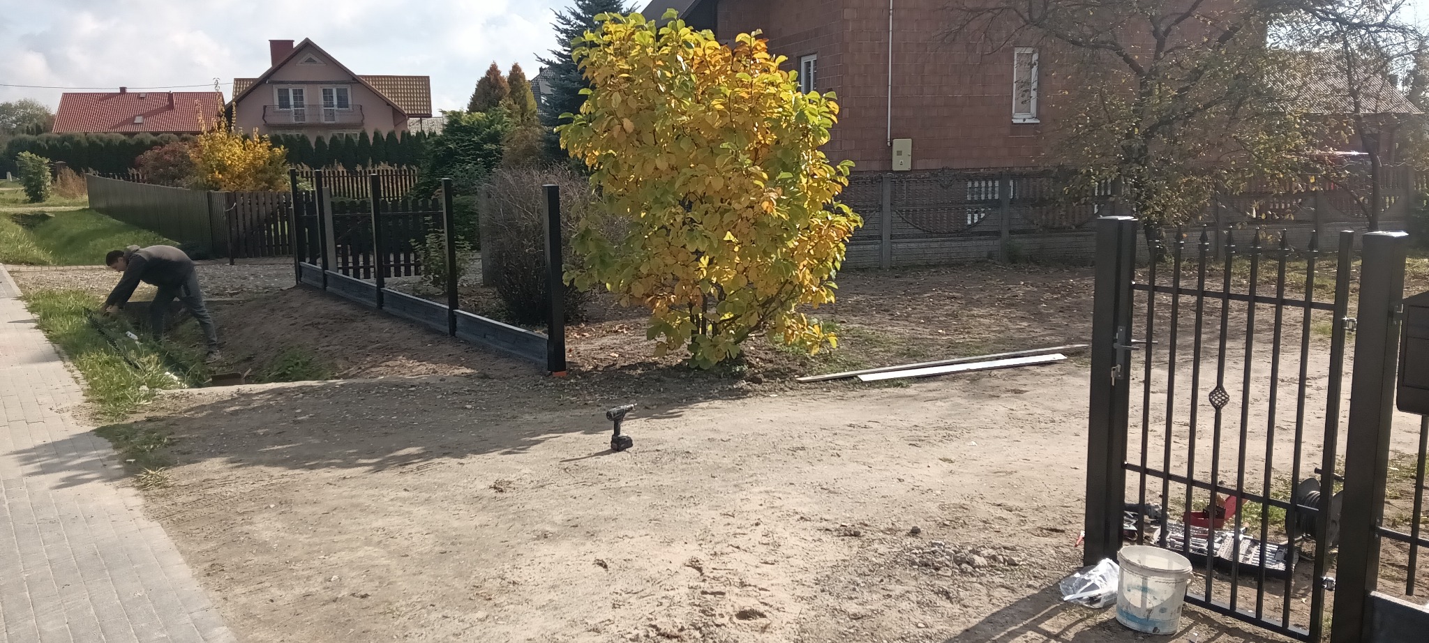 Montaż ogrodzenia panelowego: widoczny fragment ogrodzenia z czarnych paneli, narzędzia, pracownik przy wykopie. Jesienne otoczenie z drzewami i domami w tle.