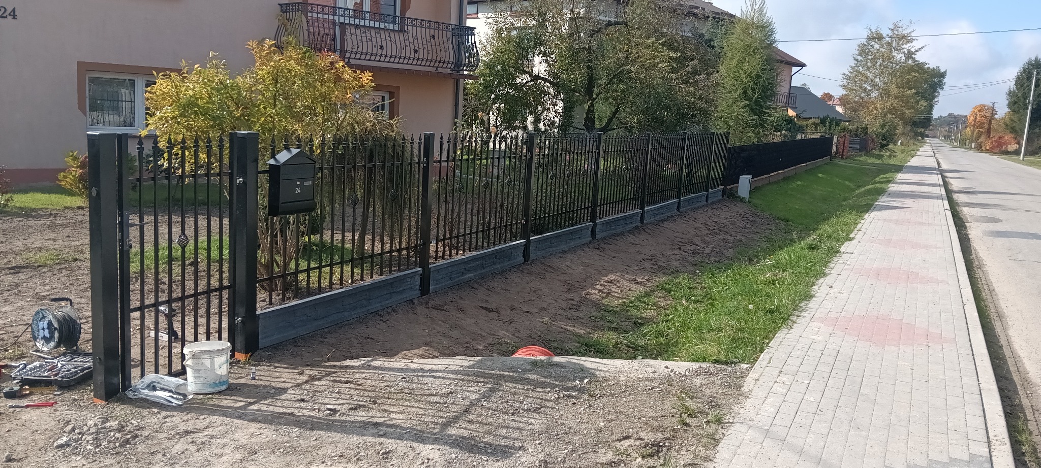 Czarne, metalowe ogrodzenie z dekoracyjnymi elementami, zamontowane na betonowej podmurówce. Widoczne narzędzia i akcesoria montażowe przy furtce.