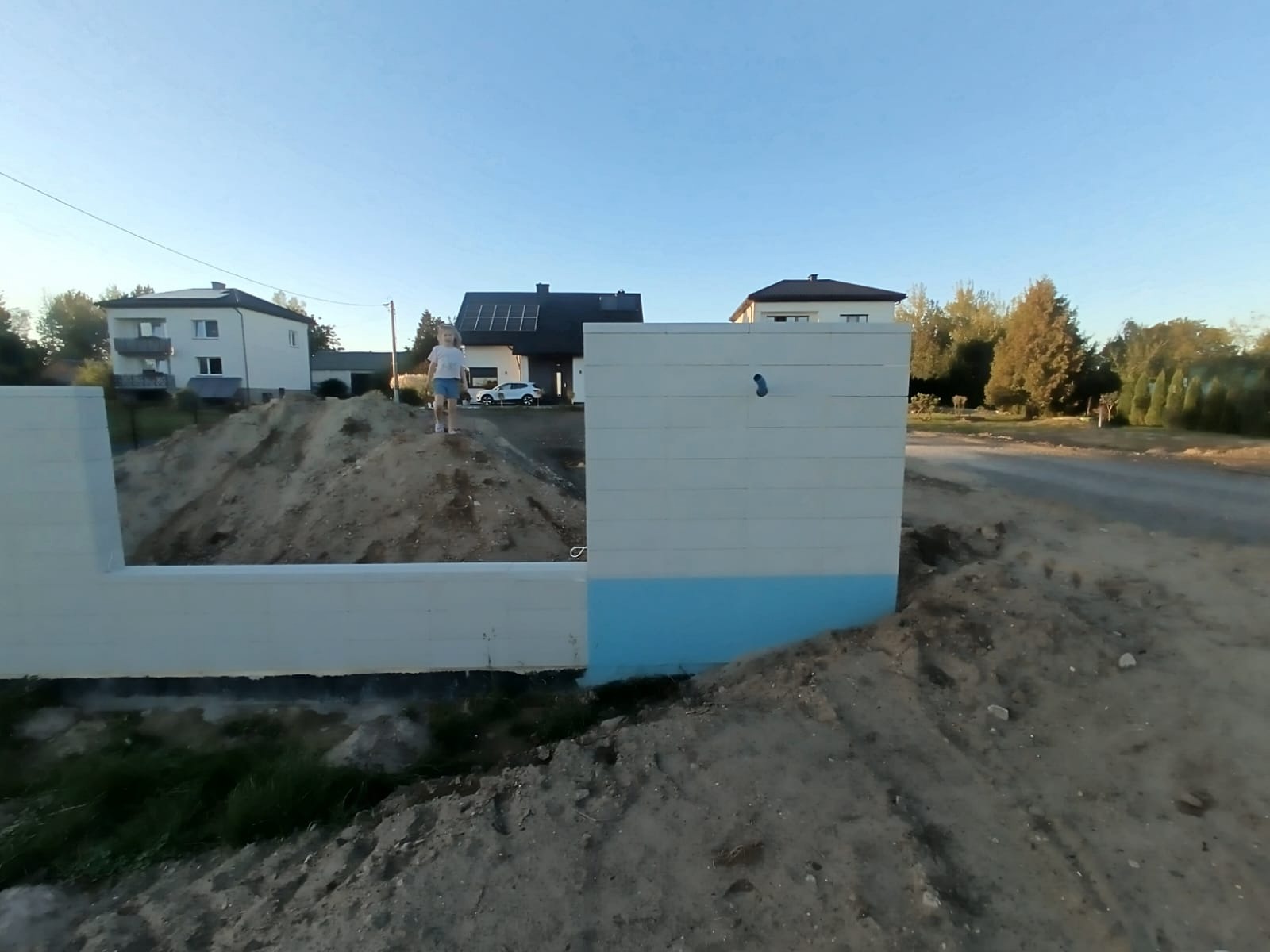 Fundament pod ogrodzenie panelowe z widocznym wykopem ziemi i osobą na górze. Tło stanowią domy i drzewa w słoneczny dzień.