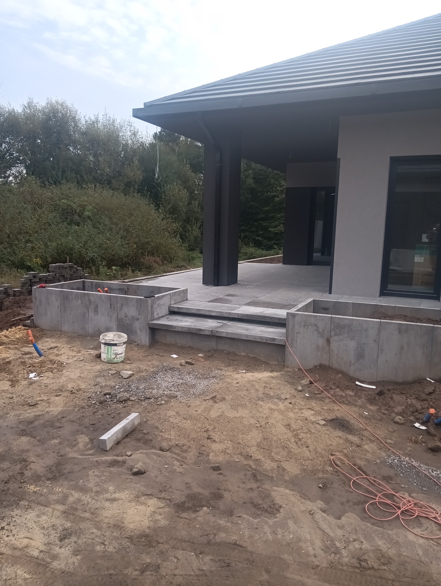 Betonowe schody i donice w trakcie budowy przed nowoczesnym domem. Widoczne materiały budowlane i narzędzia, teren przygotowany pod dalsze prace.