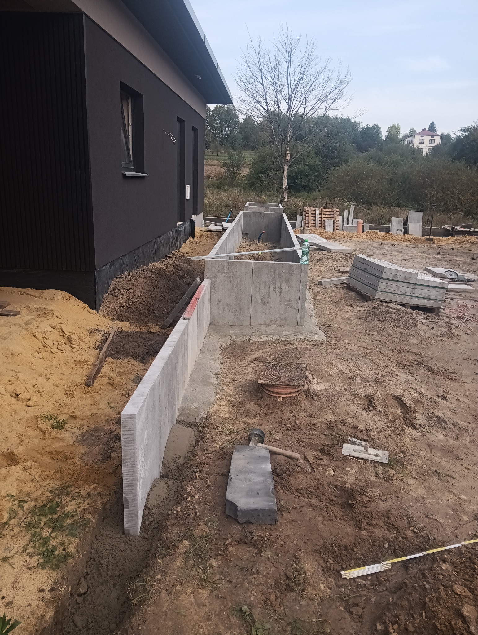 Budowa fundamentów pod ogrodzenie panelowe z betonowych prefabrykatów. Widoczne narzędzia i materiały budowlane, dom w tle.