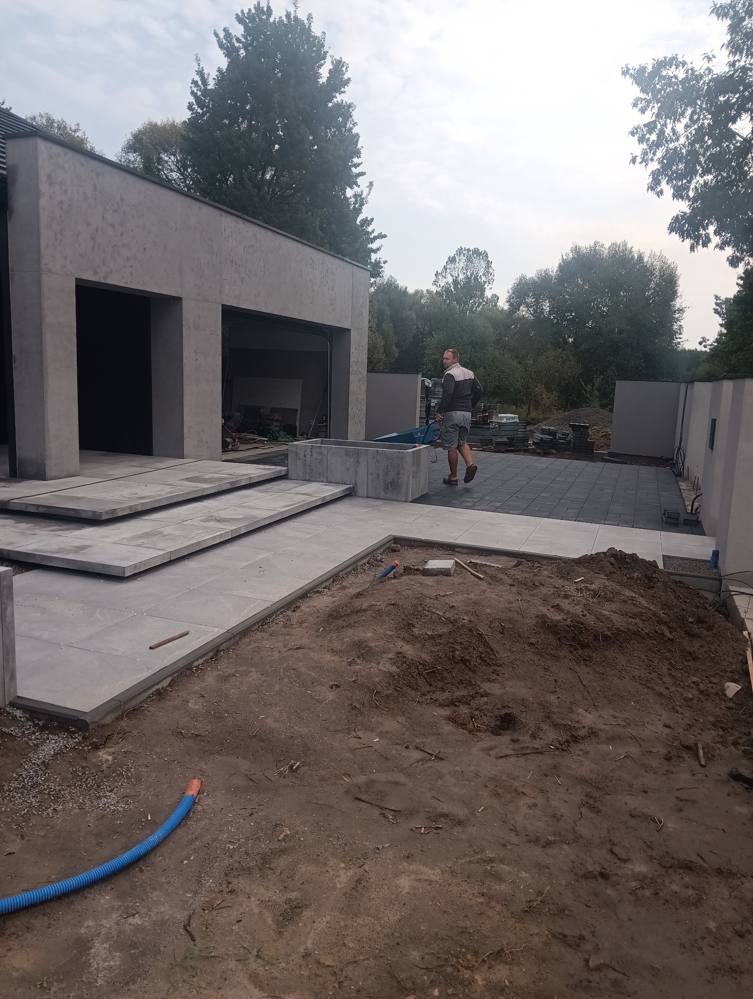 Nowoczesny dom z betonową kostką brukową na tarasie i schodach. Mężczyzna z taczką. Częściowo ułożona kostka brukowa na podwórku. Niebieski przewód w ziemi.