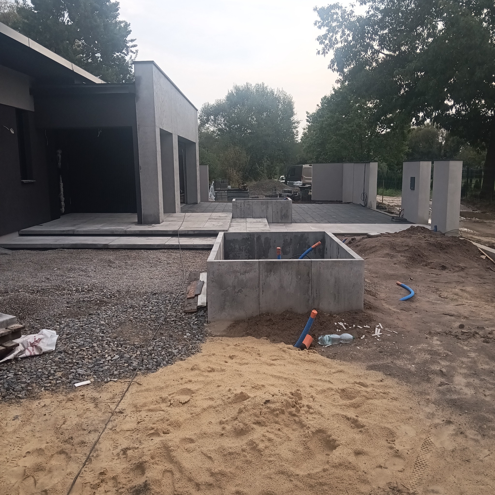 Betonowe elementy małej architektury w trakcie budowy, widoczne rury instalacyjne. W tle budynek z elementami wykończonymi kostką. Piasek na pierwszym planie.
