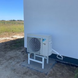 Climarte - Biała jednostka zewnętrzna klimatyzacji Daikin na wsporniku, zamontowana przy ścianie budynku. Widoczne przewody i polne tło.