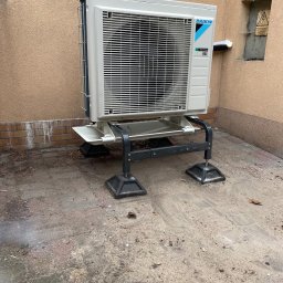 Climarte - Biała jednostka zewnętrzna klimatyzacji Daikin zamontowana na metalowej konstrukcji przy ścianie budynku, widoczny fragment instalacji elektrycznej.