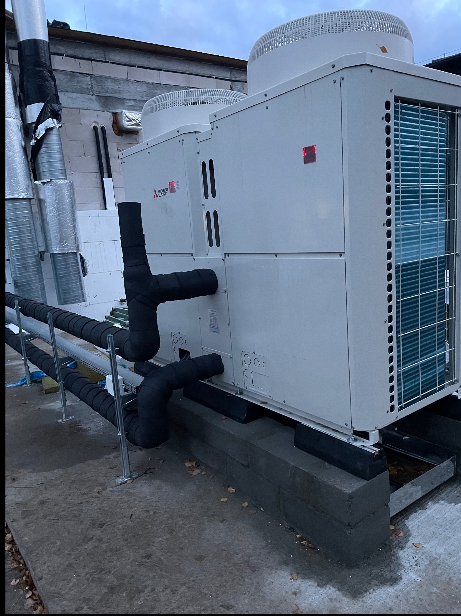 Biała jednostka zewnętrzna Mitsubishi Electric na betonowej podstawie, z widocznymi rurami w czarnej izolacji termicznej, na tle szarego nieba i fragmentu budynku.