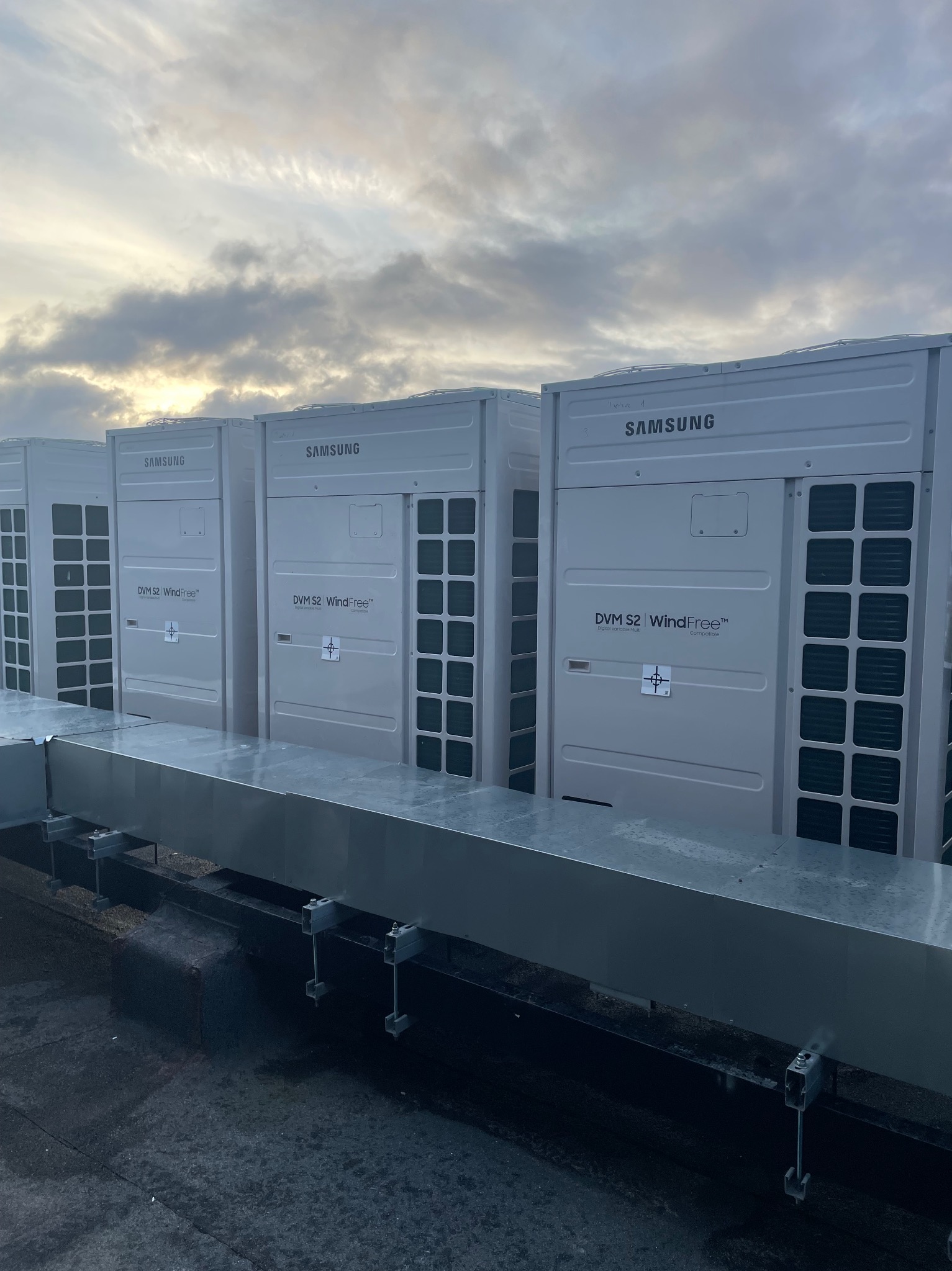 Trzy jednostki zewnętrzne klimatyzacji Samsung DVM S2 WindFree na dachu, z widocznym systemem kanałów wentylacyjnych i pochmurnym niebem w tle.