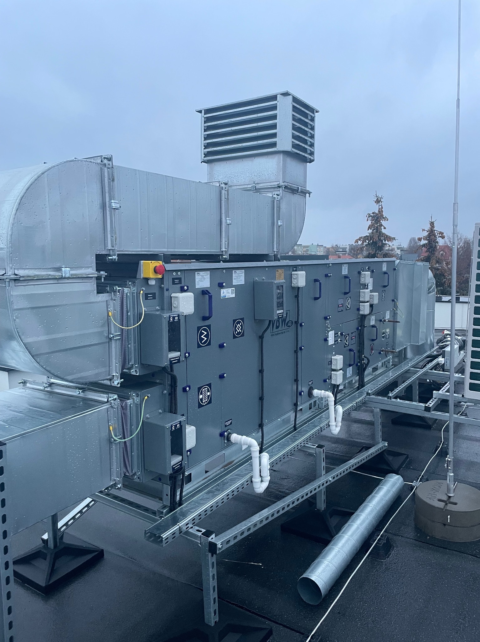 Szara centrala wentylacyjna VBW Engineering na dachu budynku, widoczna instalacja, rury, i kratki wentylacyjne. Tło to zabudowa miejska i pochmurne niebo.