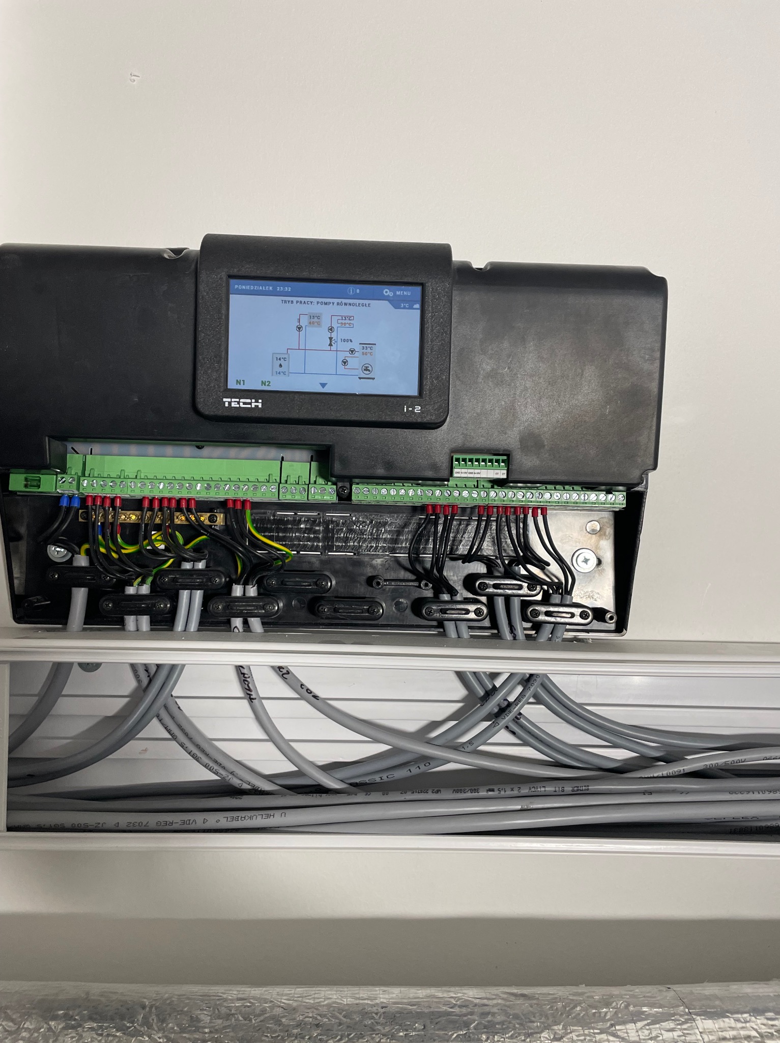 Czarny panel sterujący TECH z ekranem LCD, widoczne przewody i złącza, instalacja w białej obudowie. Kontroler pompy ciepła z widocznym schematem pracy.