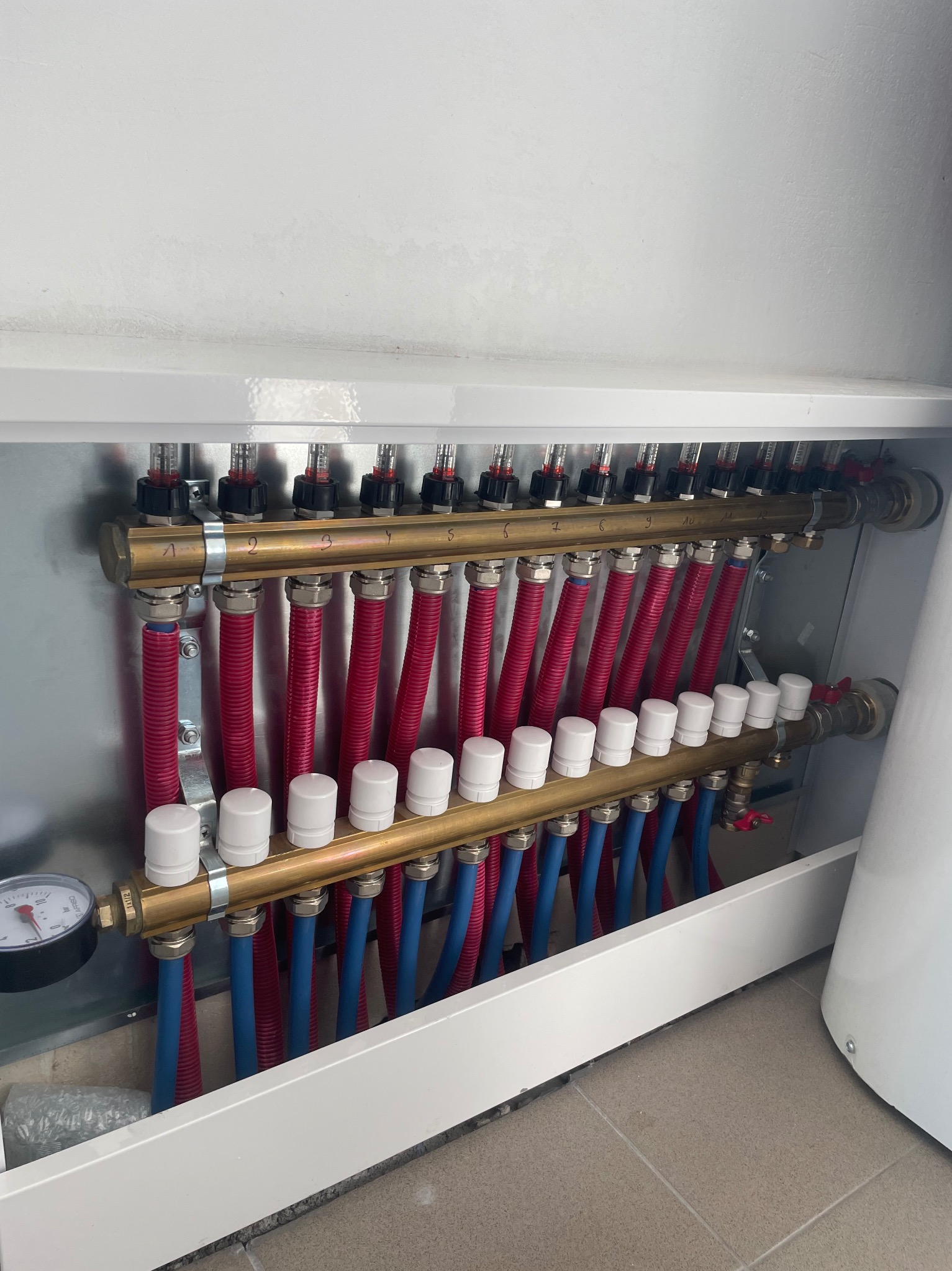 Rozdzielacz ogrzewania podłogowego z rurami w kolorach czerwonym i niebieskim, umieszczony w białej szafce z widocznym manometrem po lewej stronie.