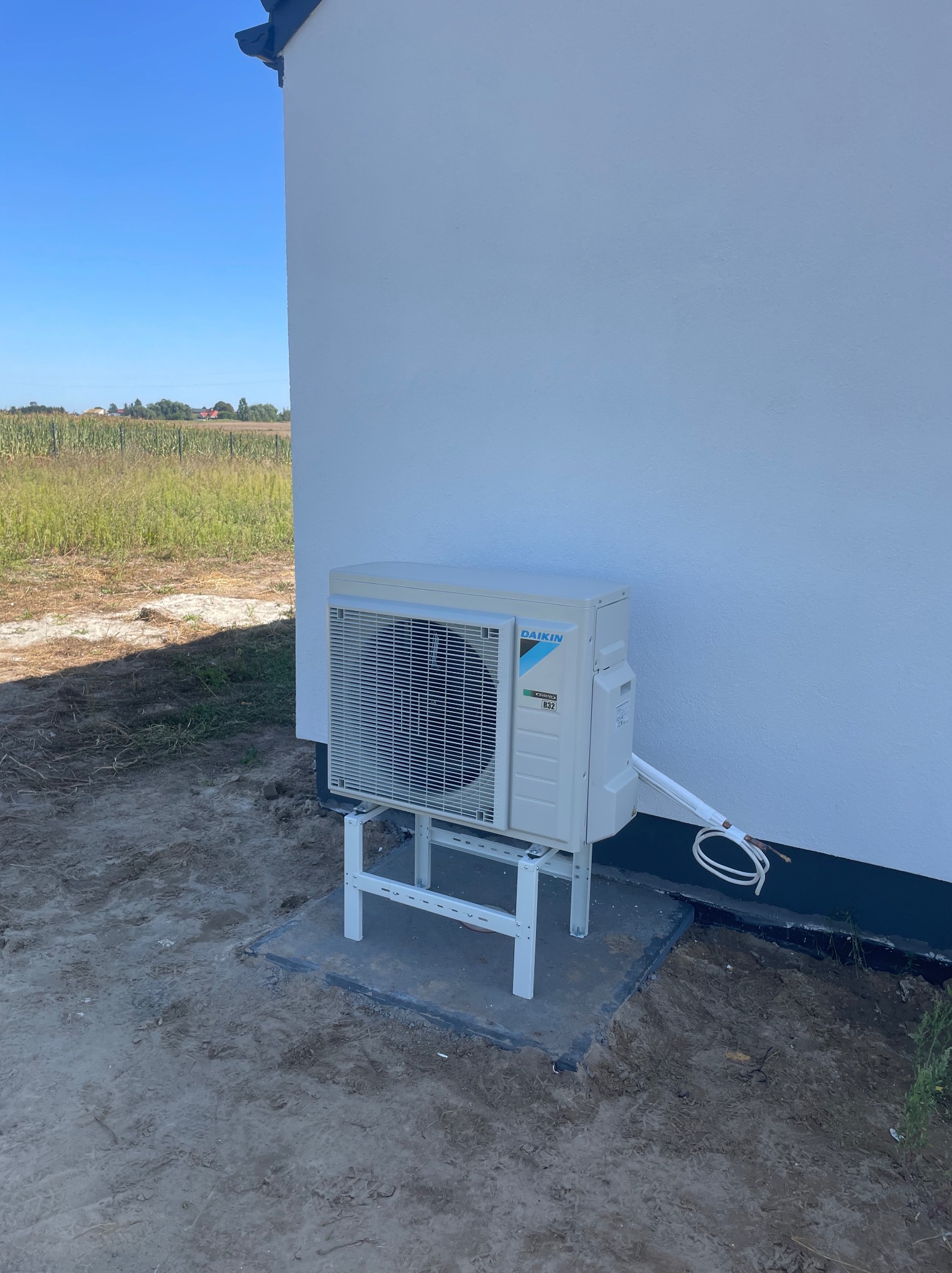 Biała jednostka zewnętrzna klimatyzacji Daikin na wsporniku, zamontowana przy ścianie budynku. Widoczne przewody i polne tło.