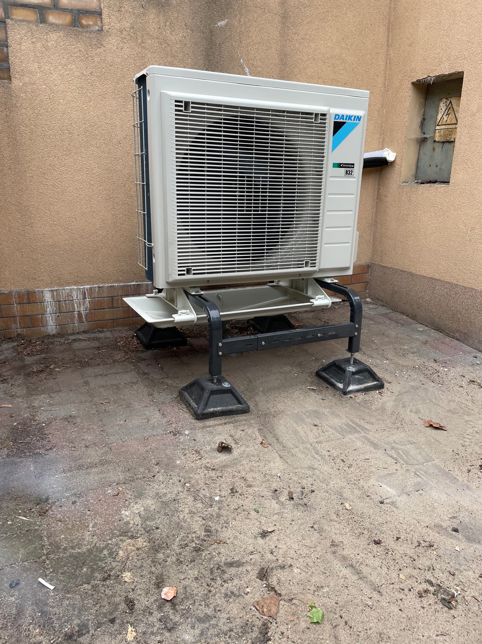 Biała jednostka zewnętrzna klimatyzacji Daikin zamontowana na metalowej konstrukcji przy ścianie budynku, widoczny fragment instalacji elektrycznej.