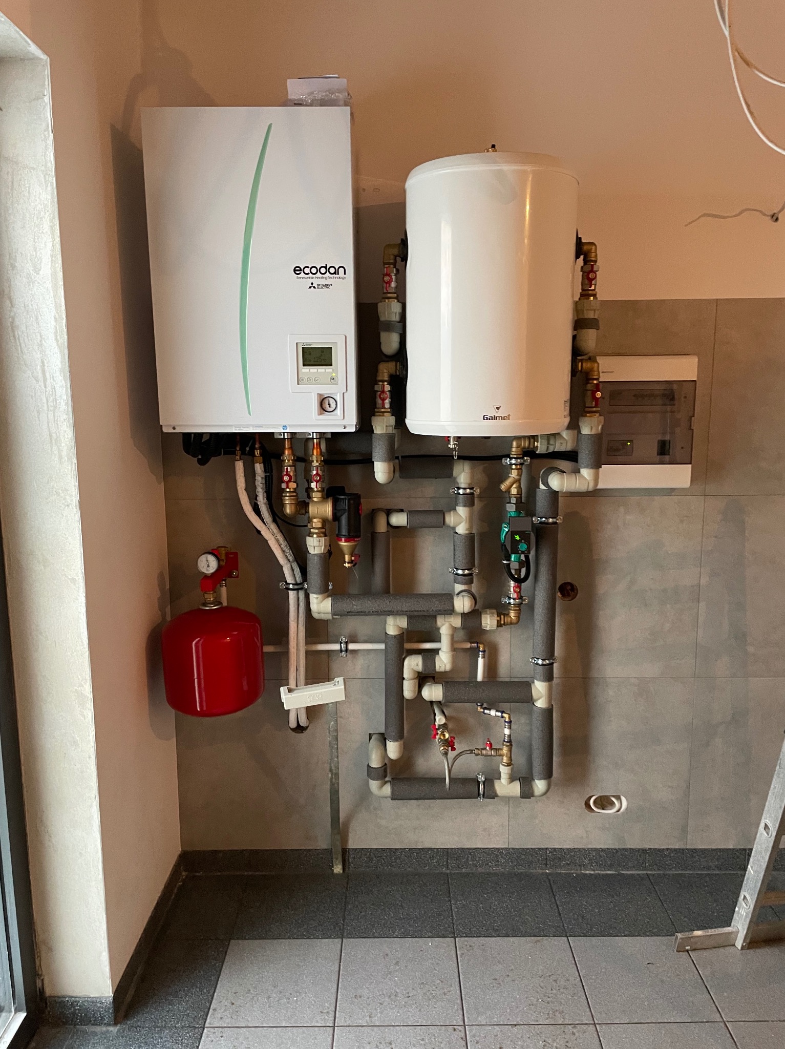 Zmontowana instalacja pompy ciepła Mitsubishi Ecodan z zasobnikiem Galmet na tle szarej ściany. Widoczne rury, zawory i czerwony zbiornik wyrównawczy.