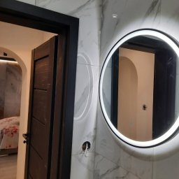 GKM Gracjan Kaźmierski - Nowoczesna łazienka z marmurowymi płytkami, okrągłym lustrem z podświetleniem LED i ciemnymi drzwiami. Widoczne niedokończone podłączenie elektryczne.