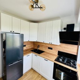 GKM Gracjan Kaźmierski - Nowoczesna kuchnia w bieli i drewnie: białe szafki z czarnymi uchwytami, drewniany blat, czarny zlew i sprzęt AGD. Lampa sufitowa z trzema szklanymi kloszami.