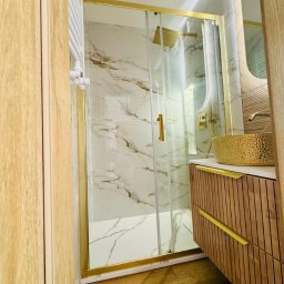 GKM Gracjan Kaźmierski - Wykończona łazienka z kabiną prysznicową w złotych ramach, marmurowe płytki, drewniana szafka z umywalką w kolorze złota. Nowoczesny design.
