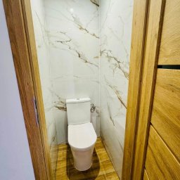 GKM Gracjan Kaźmierski - Kompaktowa łazienka z toaletą w odcieniach bieli i beżu. Marmurowe płytki ścienne z delikatnym żyłowaniem, drewnopodobna podłoga, widok zza drewnianej ościeżnicy.