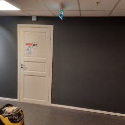 GKM Gracjan Kaźmierski - Świeżo pomalowane, ciemnoszare ściany w korytarzu z białą lamperią i drzwiami do toalety. Na podłodze widoczny odkurzacz przemysłowy.