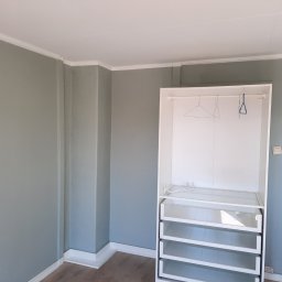 GKM Gracjan Kaźmierski - Wnętrze pokoju z szarą ścianą i białym, częściowo zmontowanym regałem z półkami i wieszakami. Widoczne listwy przypodłogowe i sufitowe.