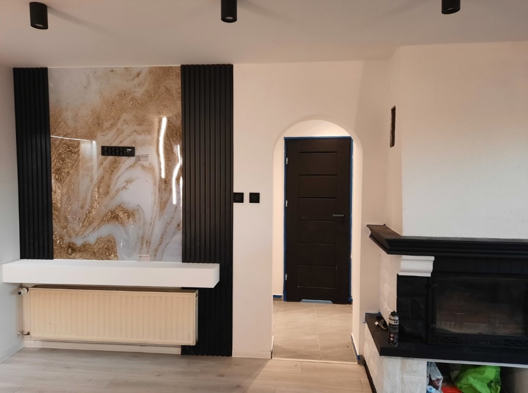 Salon po metamorfozie: ściana z dekoracyjnym panelem imitującym marmur, czarne lamele, biały kominek i ciemne drzwi w łukowatym przejściu.
