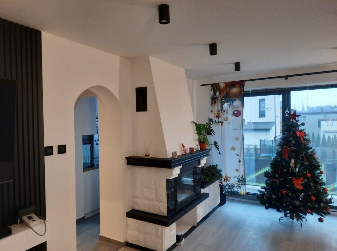 Salon po remoncie z kominkiem w stylu retro, oświetleniem punktowym i dekoracjami świątecznymi. Widok na kuchnię przez łukowe przejście.