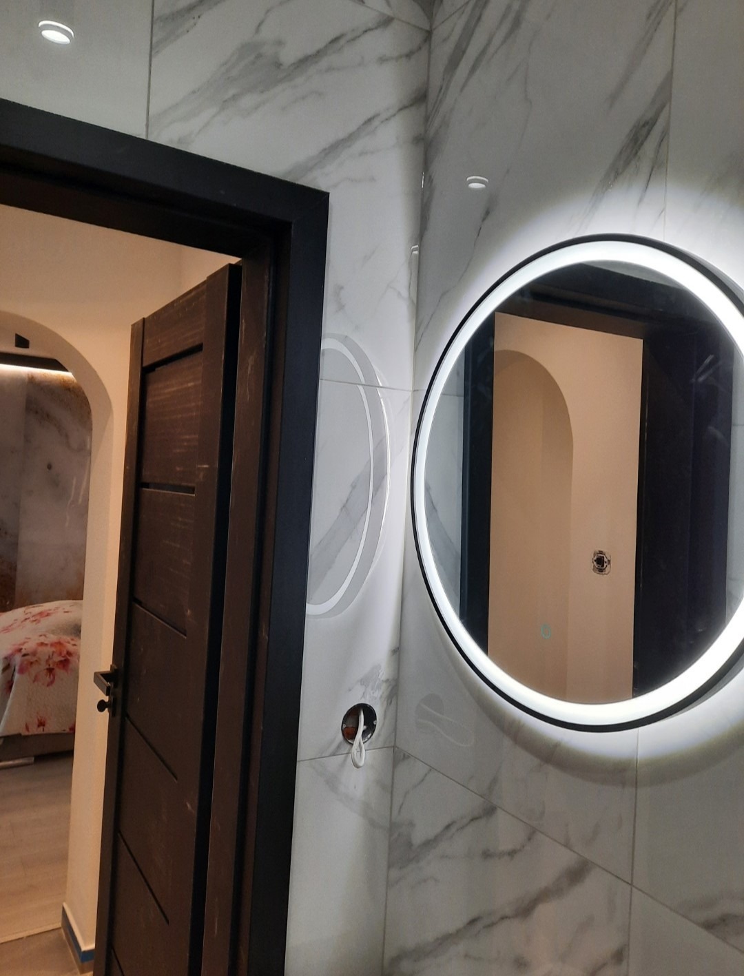 Nowoczesna łazienka z marmurowymi płytkami, okrągłym lustrem z podświetleniem LED i ciemnymi drzwiami. Widoczne niedokończone podłączenie elektryczne.