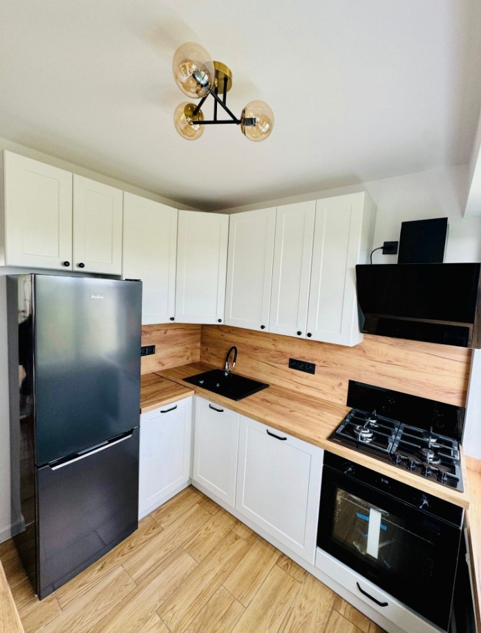 Nowoczesna kuchnia w bieli i drewnie: białe szafki z czarnymi uchwytami, drewniany blat, czarny zlew i sprzęt AGD. Lampa sufitowa z trzema szklanymi kloszami.