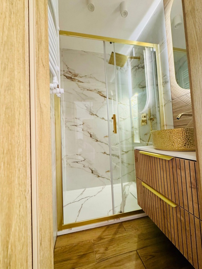 Wykończona łazienka z kabiną prysznicową w złotych ramach, marmurowe płytki, drewniana szafka z umywalką w kolorze złota. Nowoczesny design.