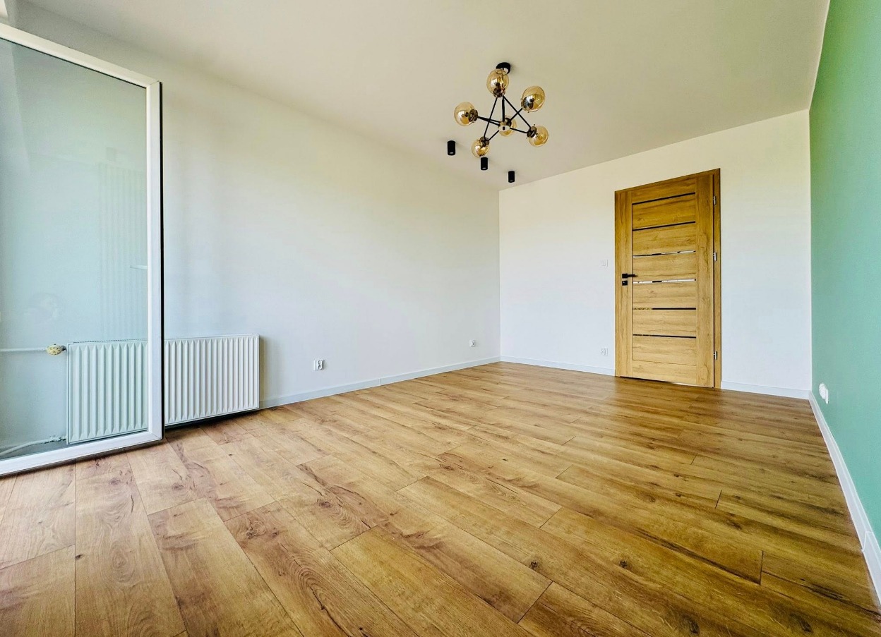 Jasne wnętrze z drewnianą podłogą, nowoczesną lampą i drzwiami. Ściany białe, jedna zielona. Widoczny grzejnik i okno. Minimalistyczny design.