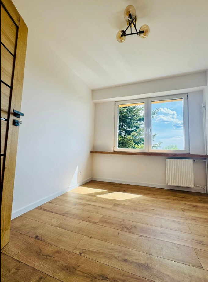 Jasne wnętrze pokoju po remoncie: drewniana podłoga, białe ściany, okno z widokiem na zieleń, nowoczesna lampa sufitowa i drewniane wykończenie.