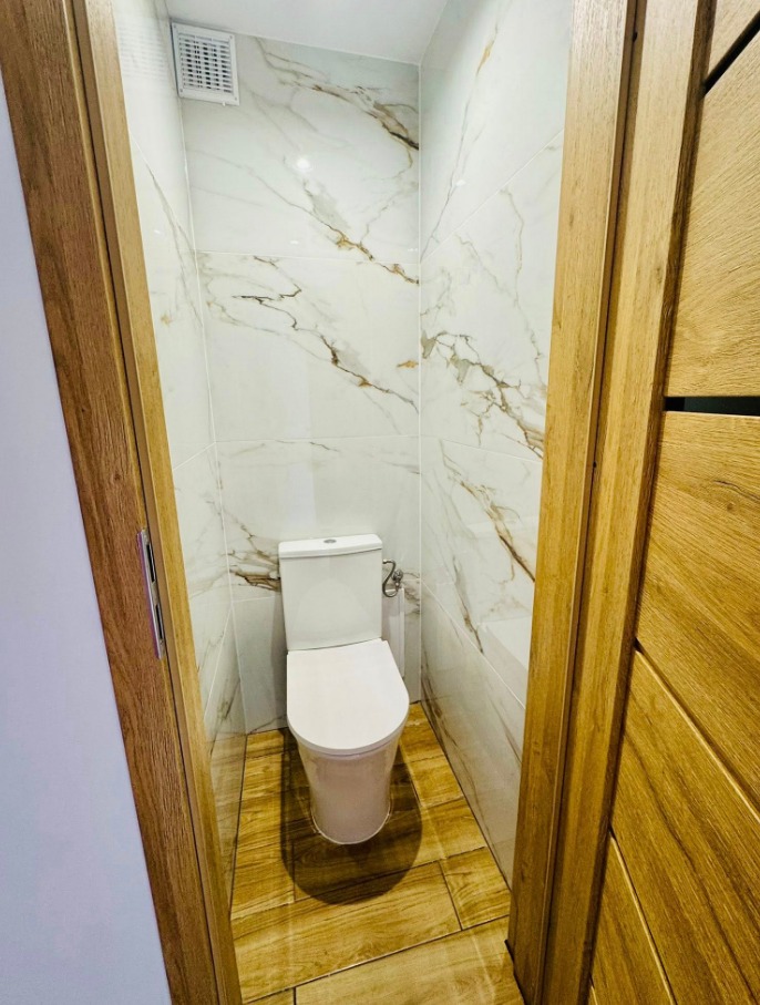 Kompaktowa łazienka z toaletą w odcieniach bieli i beżu. Marmurowe płytki ścienne z delikatnym żyłowaniem, drewnopodobna podłoga, widok zza drewnianej ościeżnicy.