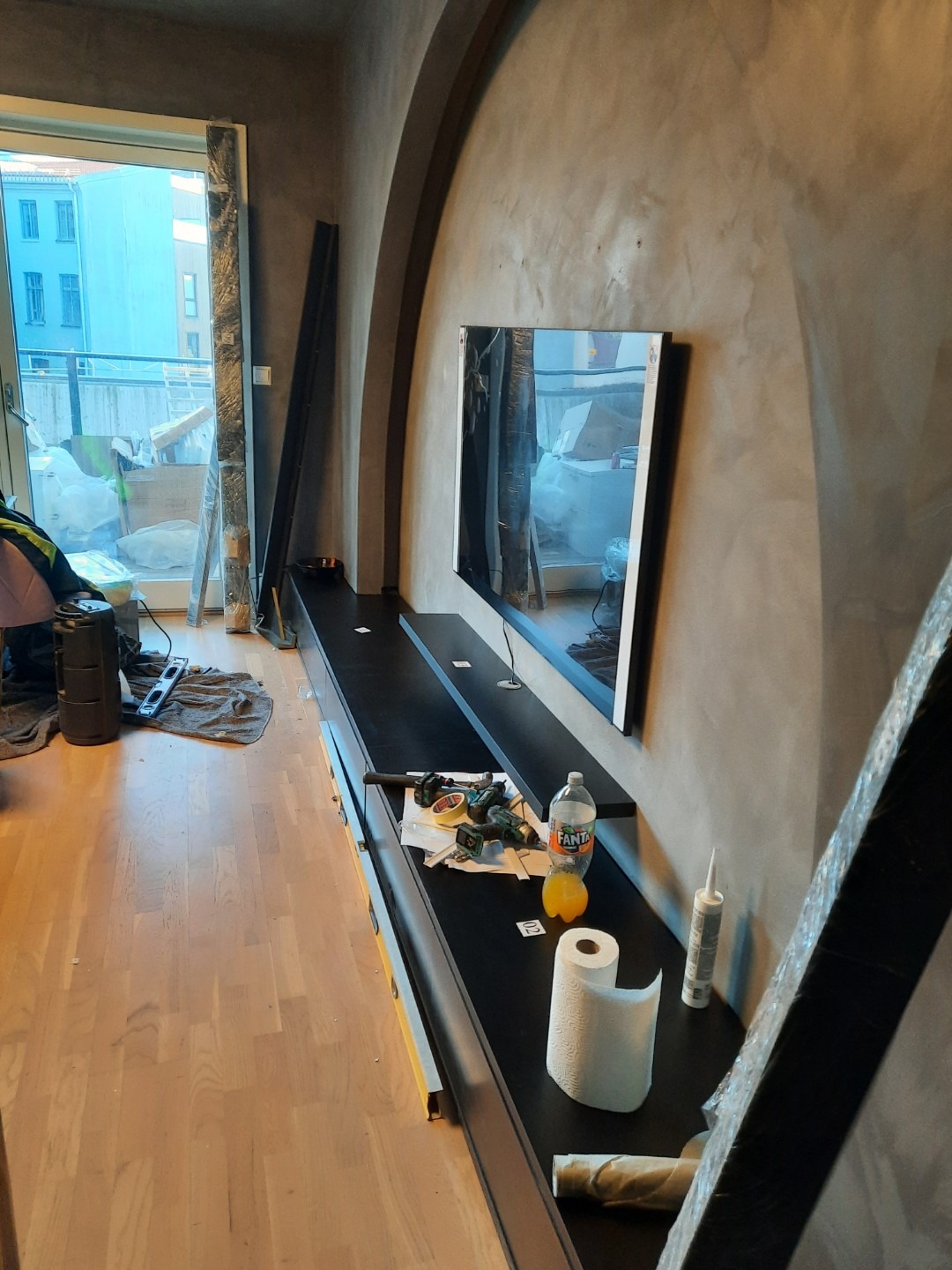 Wnętrze w trakcie prac wykończeniowych: zamontowana szafka RTV pod telewizorem, narzędzia i materiały budowlane na blacie, surowe ściany w odcieniach szarości.