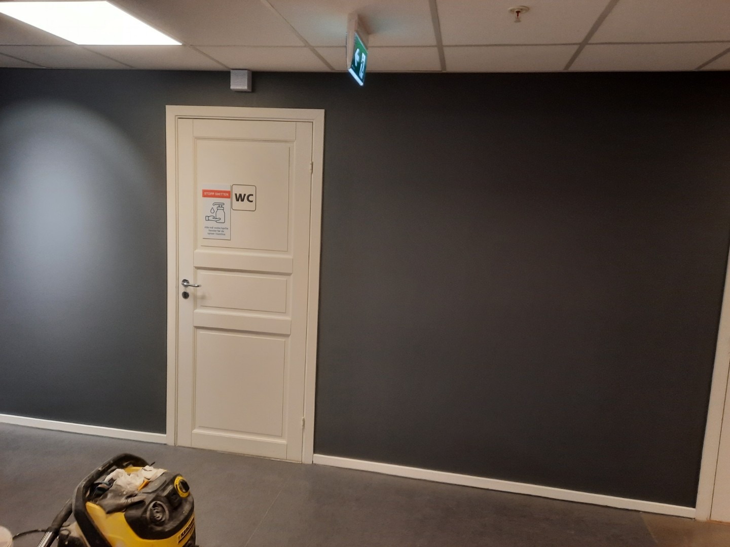 Świeżo pomalowane, ciemnoszare ściany w korytarzu z białą lamperią i drzwiami do toalety. Na podłodze widoczny odkurzacz przemysłowy.