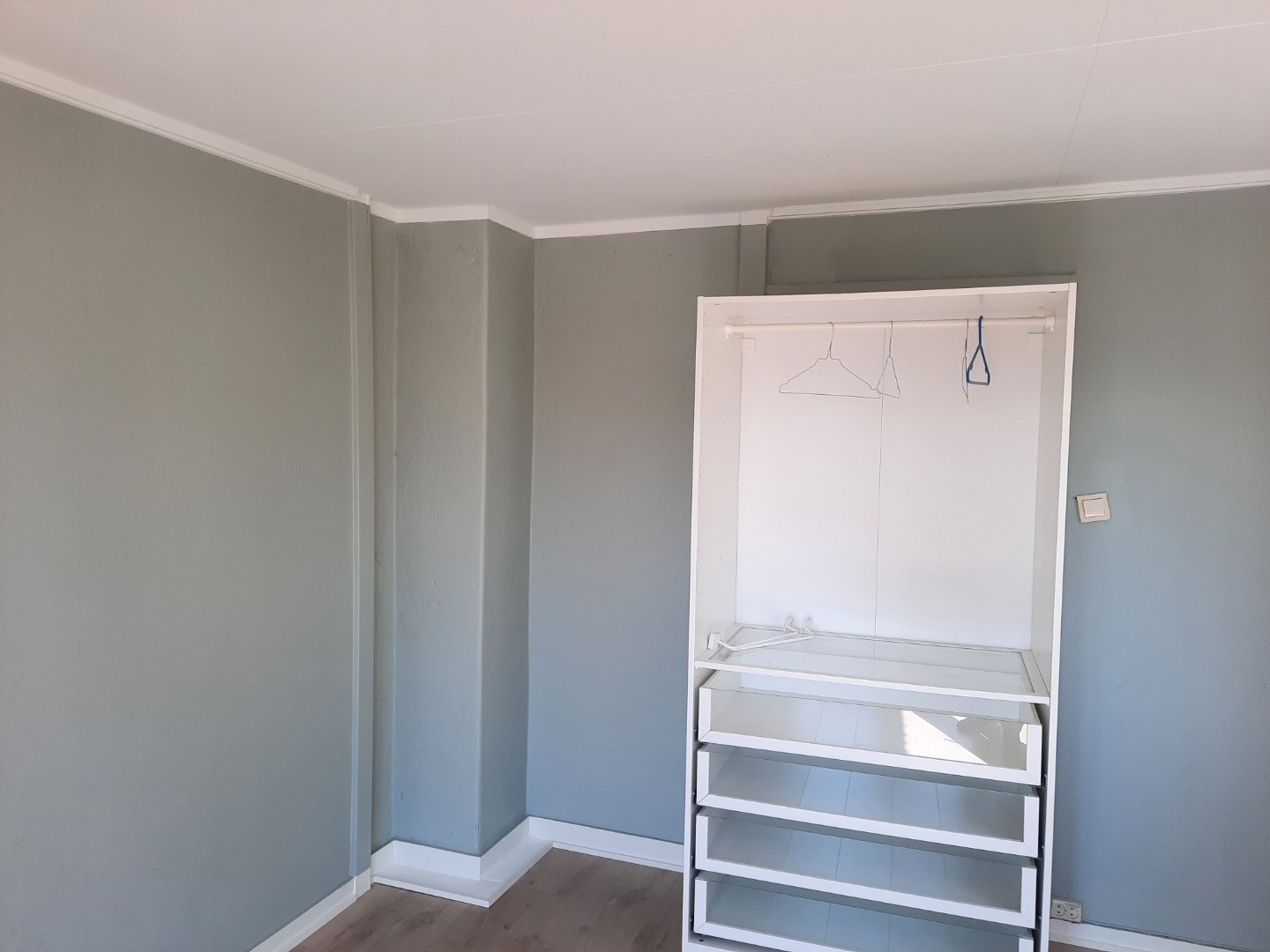 Wnętrze pokoju z szarą ścianą i białym, częściowo zmontowanym regałem z półkami i wieszakami. Widoczne listwy przypodłogowe i sufitowe.